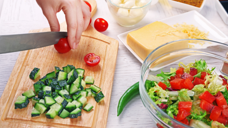 Hand cutting cherry tomatoes on a chopping board beside a salad bowl and chopped vegetables.