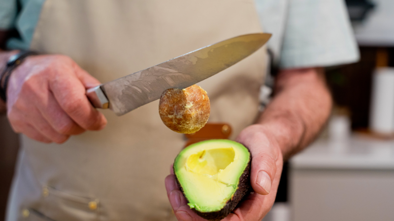 Kitchen knife being used to remove the pit from a halved avocado held in one hand.